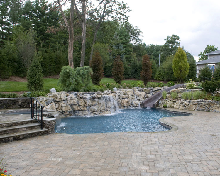Custom natural-style pool with rock waterfalls and a water slide, surrounded by greenery and a paved patio area.